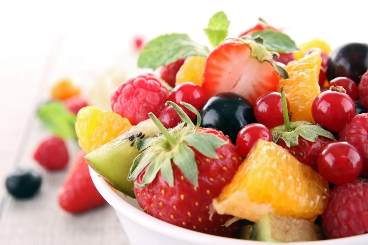 Summer fruit salad in a white bowl with a blurred background of berries and a white wooden table