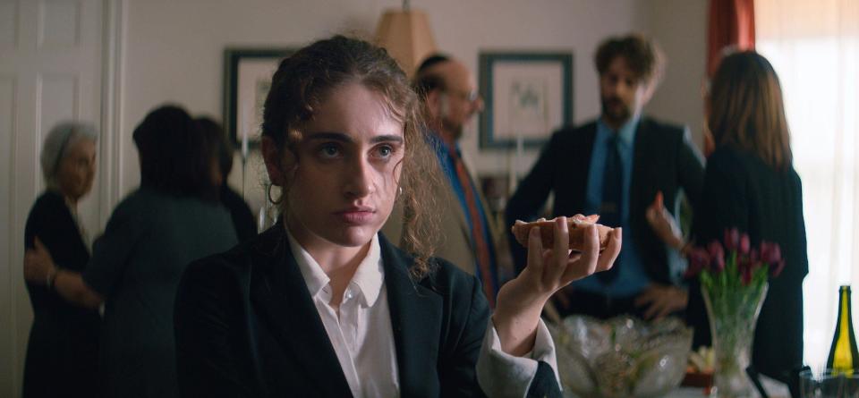 An angry woman in a suit holds up a bagel during a shiva