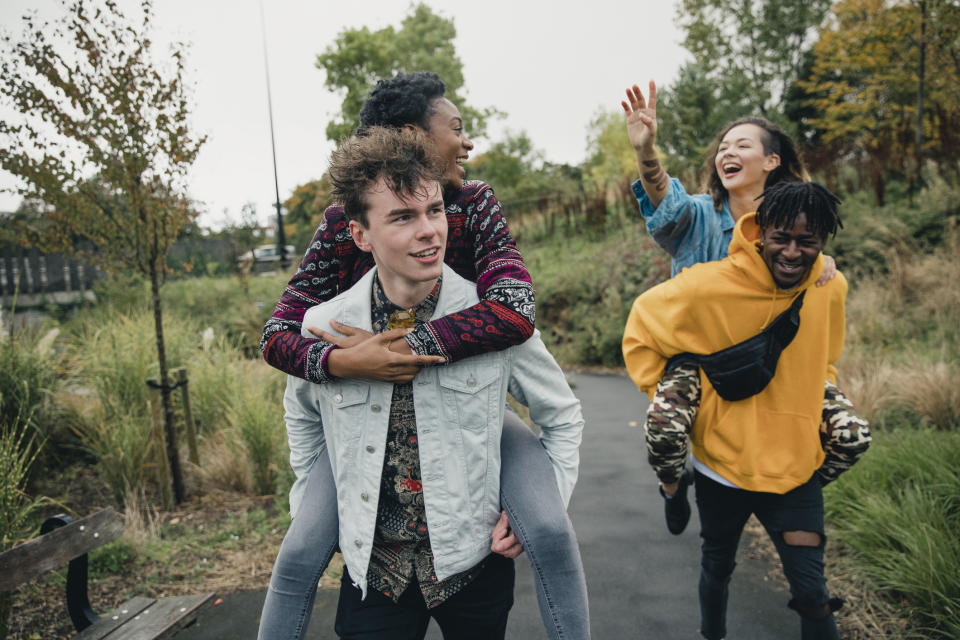 Two men giving piggyback rides to their girlfriends in a park, all smiling and enjoying the moment