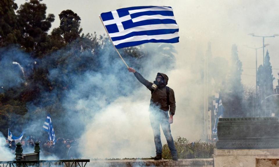 A man holds a Greek flag in a cloud of teargas.