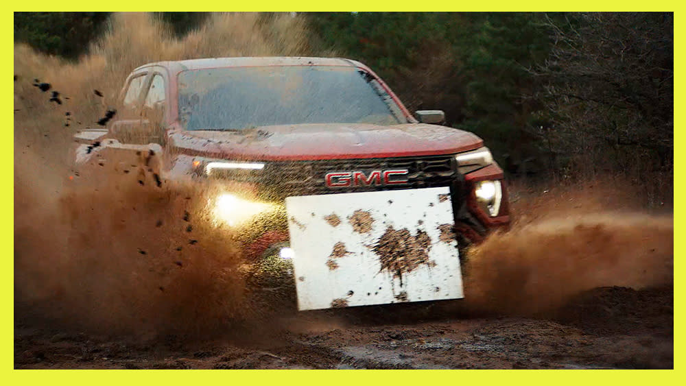  A GMC truck drives through mud with an art canvas attached to its front grille. 