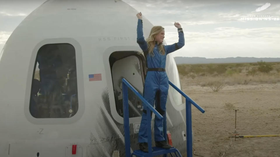  A woman in a blue flight suit raises her arms in triumph after exiting a white space capsule in the desert. 