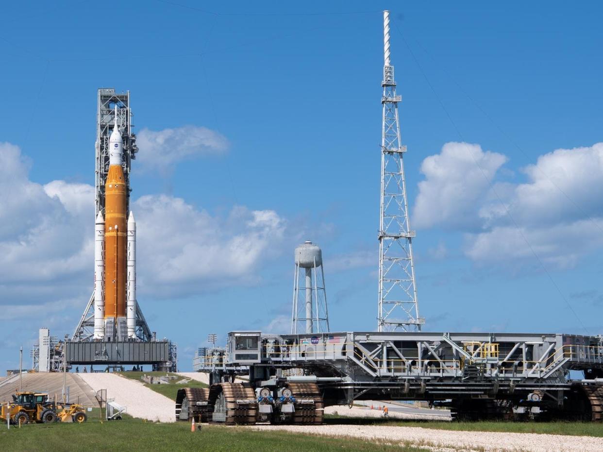 flat wide platform with wheels on a runway in front of giant orange rocket against blue sky