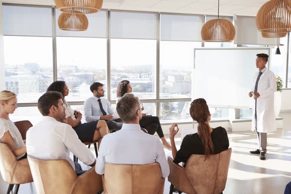 A medical professional in a white coat gives a presentation with a white board to seven spectators.