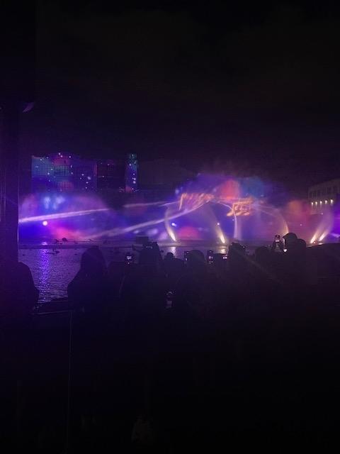 Water show in progress at night, with colorful lights and projections on water sprays. A crowd of people watch and capture the event with their cameras