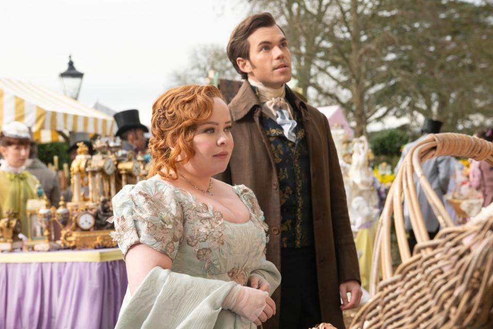 Nicola Coughlan and Luke Newton in period costumes, standing near a market stall in a historical setting, both looking ahead
