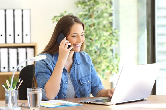 Smiling woman on phone looks at laptop on desk.