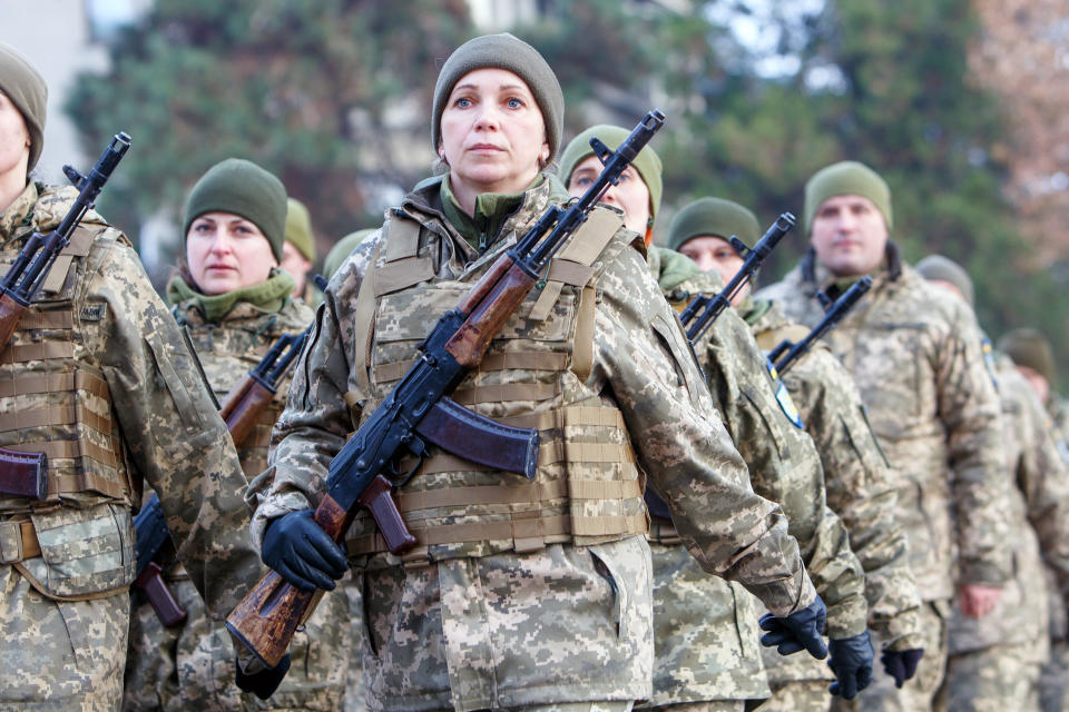 A column of male and female soldiers armed with rifles stride ahead, with trees behind.