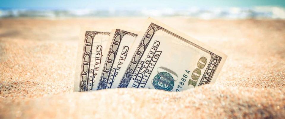 Money dolars half covered with sand lie on sandy beach near sea ocean waves on sunny summer day close-up. Money grows out of the ground. Concept finance money holiday relax vacation.