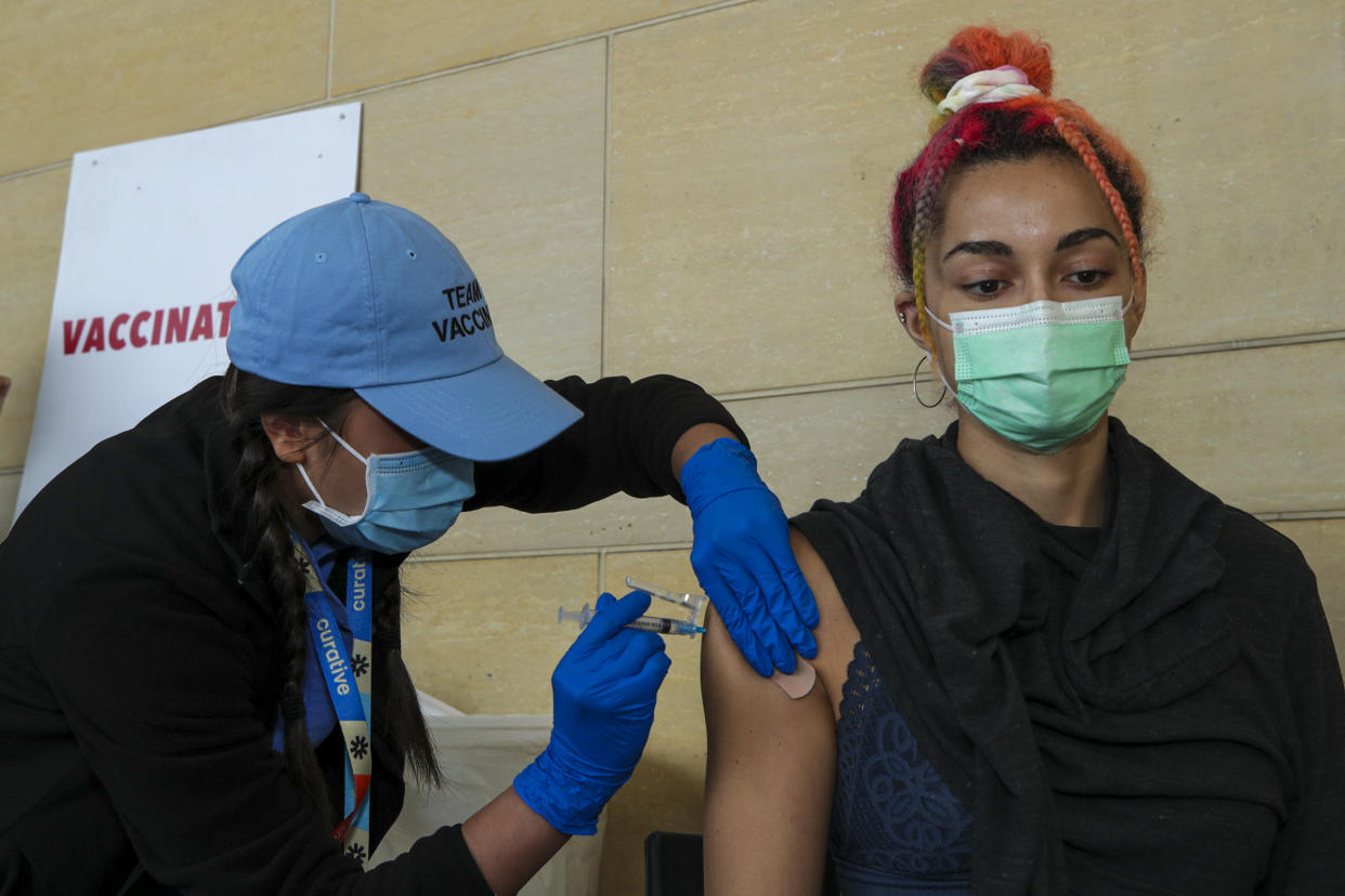 A healthcare worker wearing rubber gloves gives a vaccine shot to a woman's upper arm. Both wear face masks