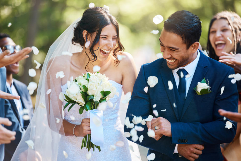 A joyful couple, bride in a white dress and groom in a blue suit, surrounded by guests throwing flower petals, celebrating their wedding day outdoors