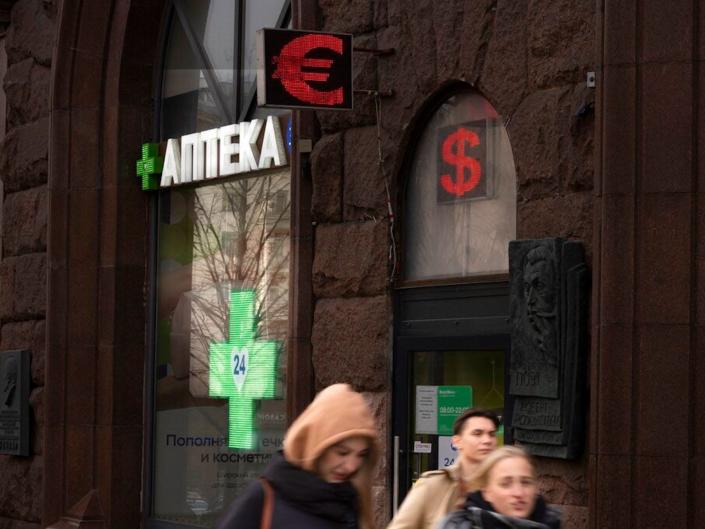 People walk past a pharmacy and a currency exchange screen in a main street in Moscow, Russia, Friday, April 1, 2022.