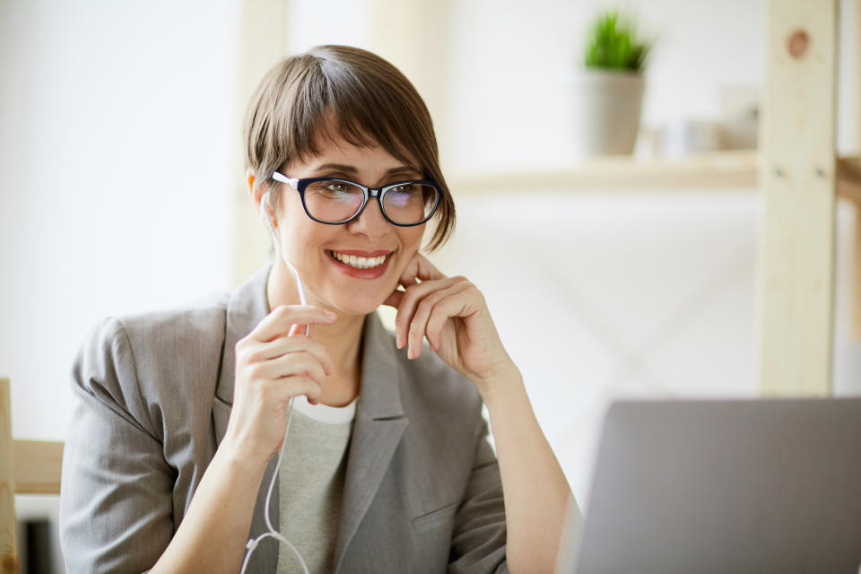 Portrait of young successful businesswoman wearing creative haircut and glasses smiling cheerfully while holding videochat using hands free mic and laptop at workplace in modern office against window