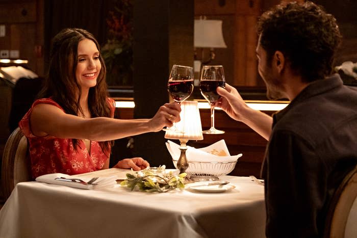 Natalie and a man make a toast with their glasses of wine as they sit in a restaurant