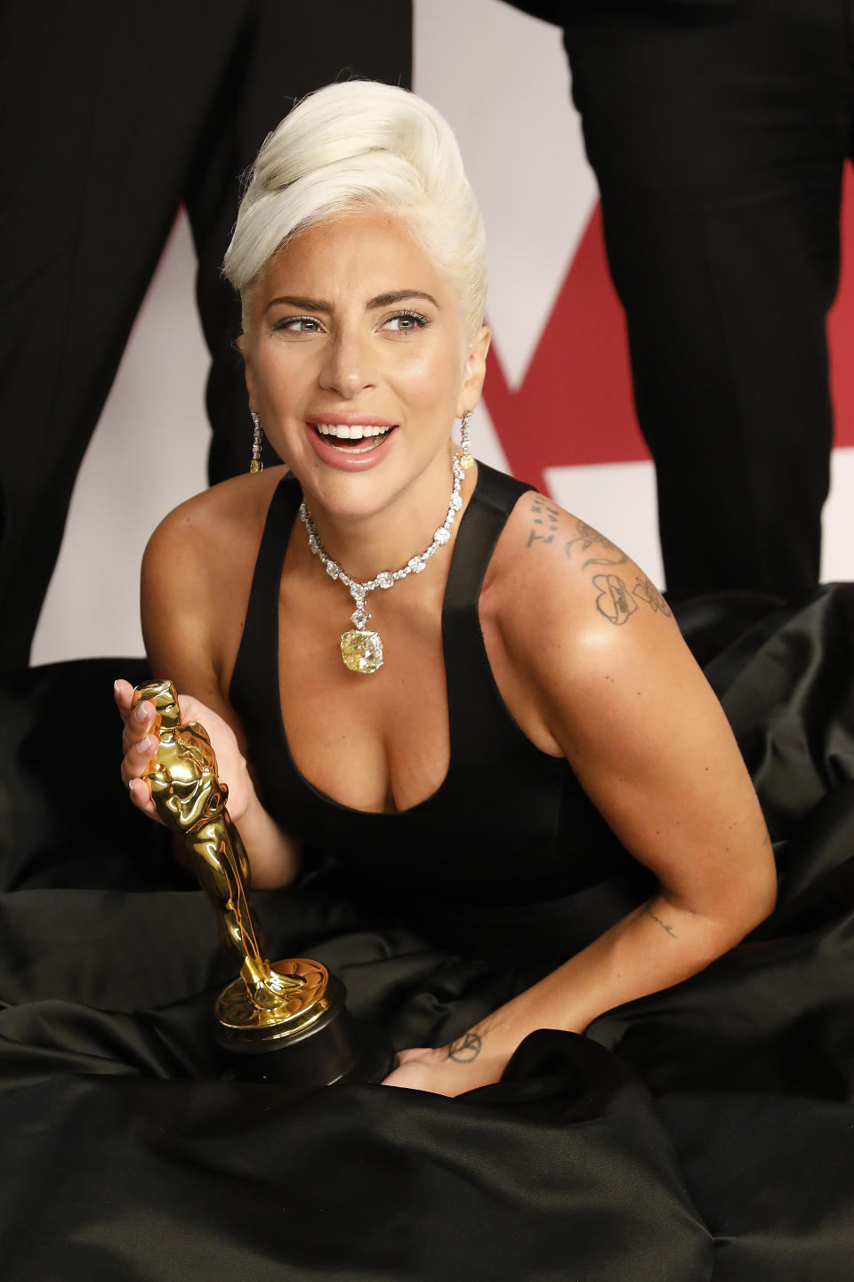 Lady Gaga holding an Oscar on a red carpet, wearing a sleek black dress and a statement diamond necklace, smiling towards the camera