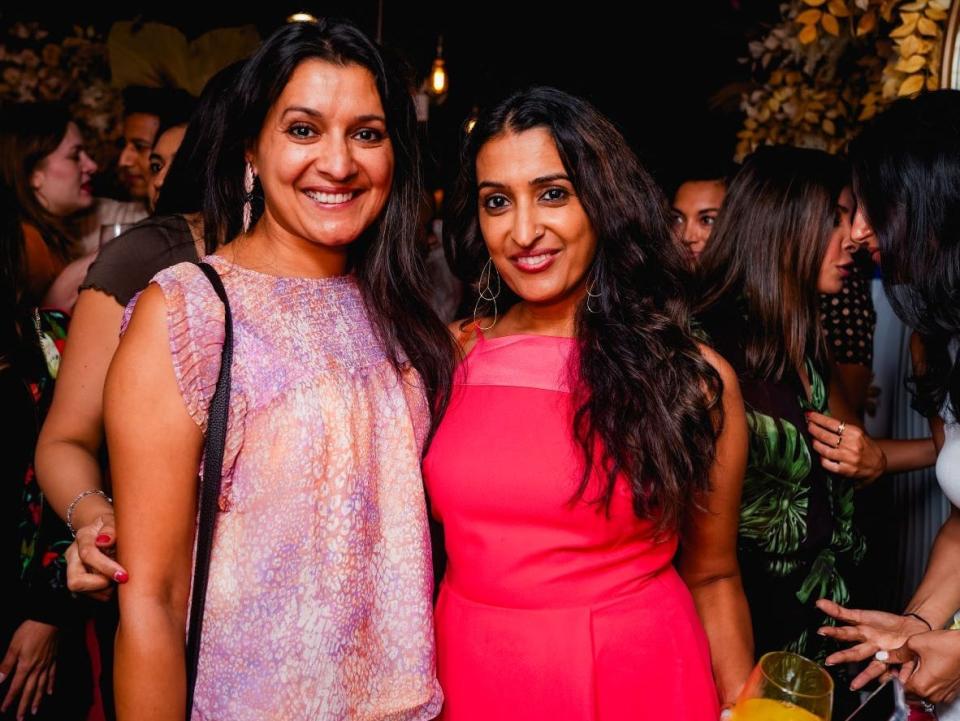 A photo of Shailja Ambrose on the left and Neerja Patel on the right. They are smiling and standing in a crowd indoors. Shailja wears a pink patterned shirt and jeans. Neerja wears a a red square-necked dress and holds a wine glass in her left hand, while the right hand is around her sister's waist.