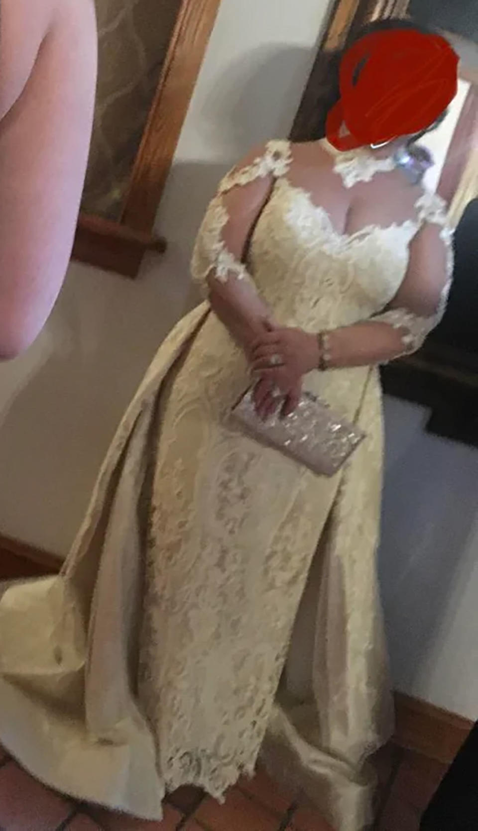 The wedding guest wearing a lace detailed floor length white gown, holding a sparkly clutch bag, her face is covered with a red dot