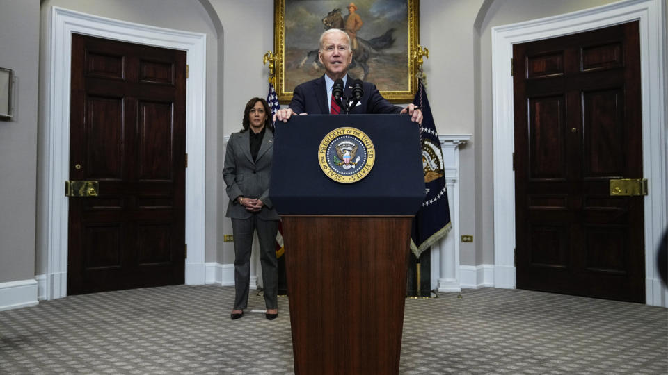 Vice President Kamala Harris stands far behind the podium where President Biden is speaking, under an equestrian portrait of President Theodore Roosevelt.