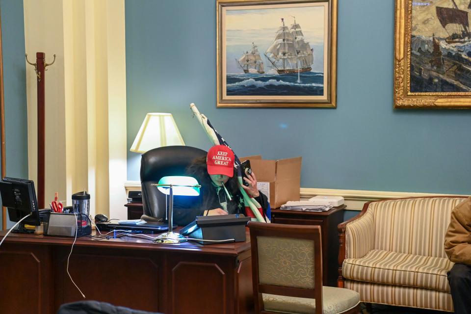 A supporter of President Donald Trump holding an American flag sits at a desk after invading the Capitol on Jan. 6 in Washington. Some wrote menacing notes in lawmakers’ offices and smoked after breaking in.