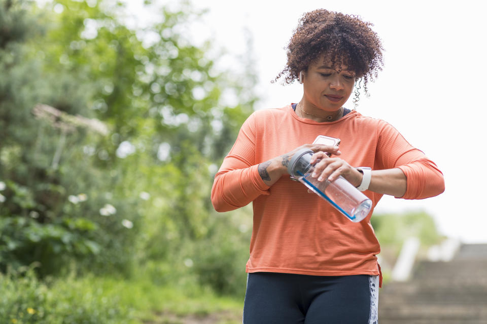 A person in athletic attire checks their smartwatch while holding a water bottle, standing outdoors with greenery and stairs in the background