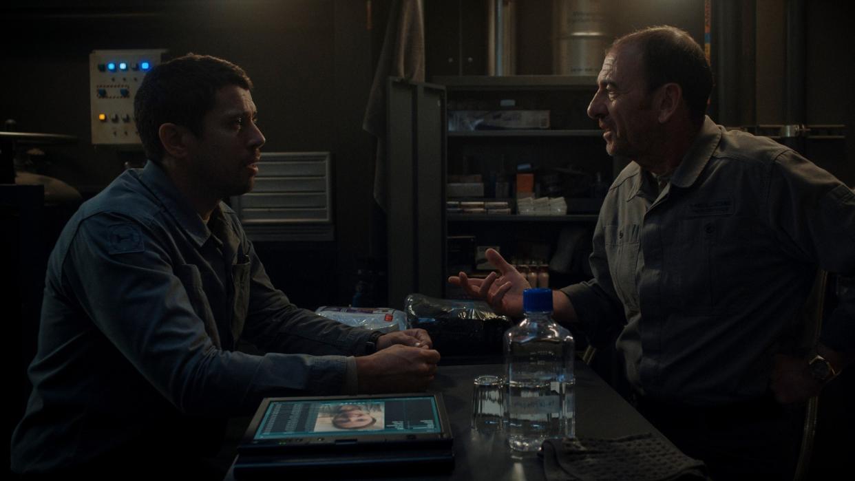  Two men sitting either side of a desk in an office. Both are wearing grayish-blue jumpsuits. The older man is explaining something to the younger man. 