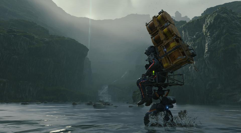 Sam making his way across a river with a huge pack on his back in Death Stranding.