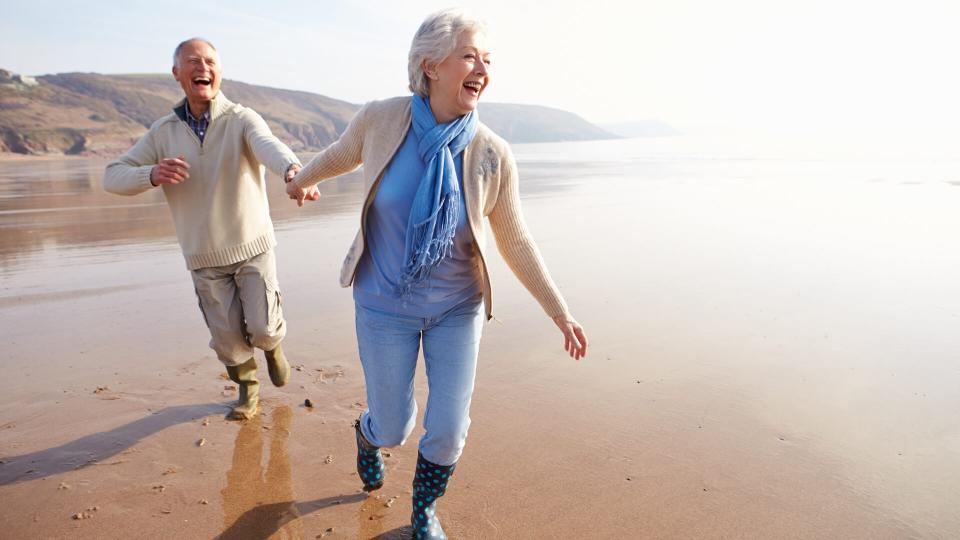 Senior Couple Running Along Winter Beach Laughing.