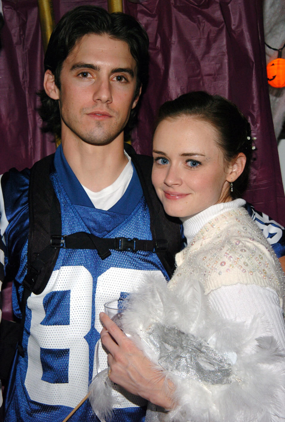 Milo Ventimiglia in a football jersey standing next to Alexis Bledel who's wearing a  sequined sweater