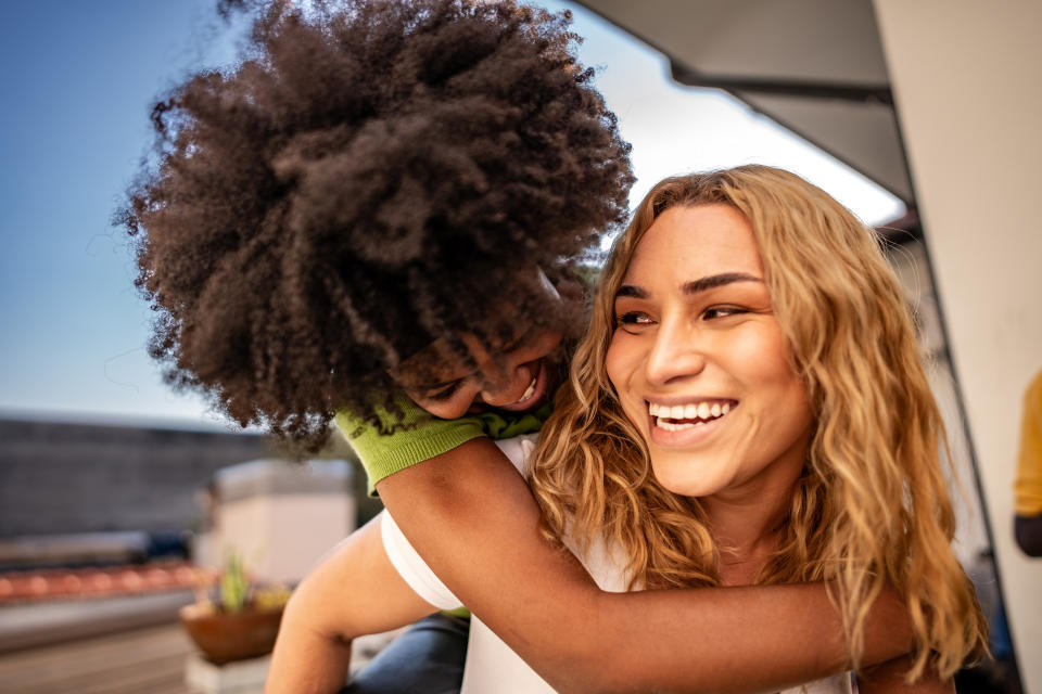 Two people laughing and hugging, one with curly hair and the other with wavy hair. Both display joy and affection in a friendly embrace