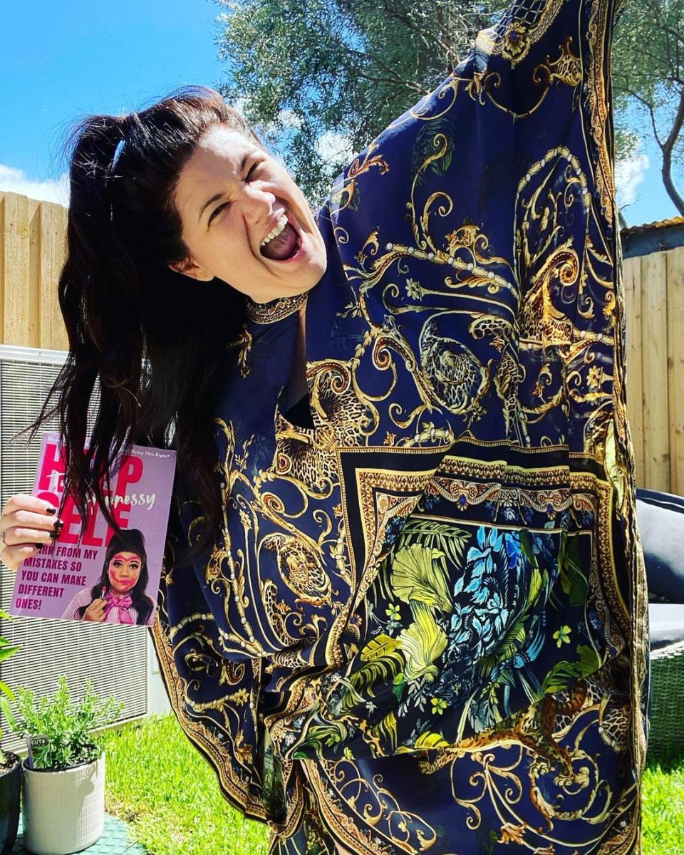 Tanya Hennessy wearing a blue and gold kaftan while holding a copy of her new book, Help Self