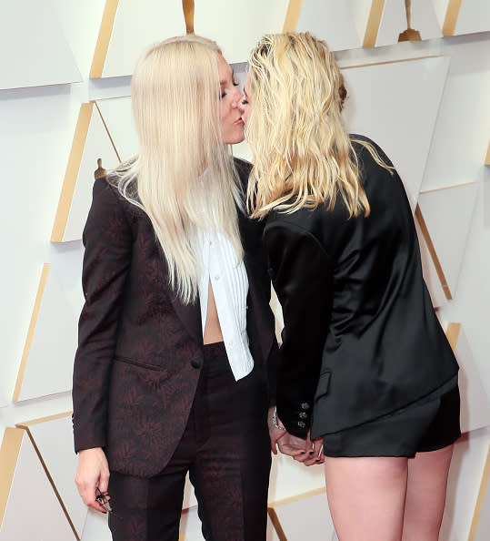 Gwyneth Paltrow and Kristen Stewart share a kiss on the red carpet while holding hands. Paltrow is in a suit, Stewart in a blazer and shorts