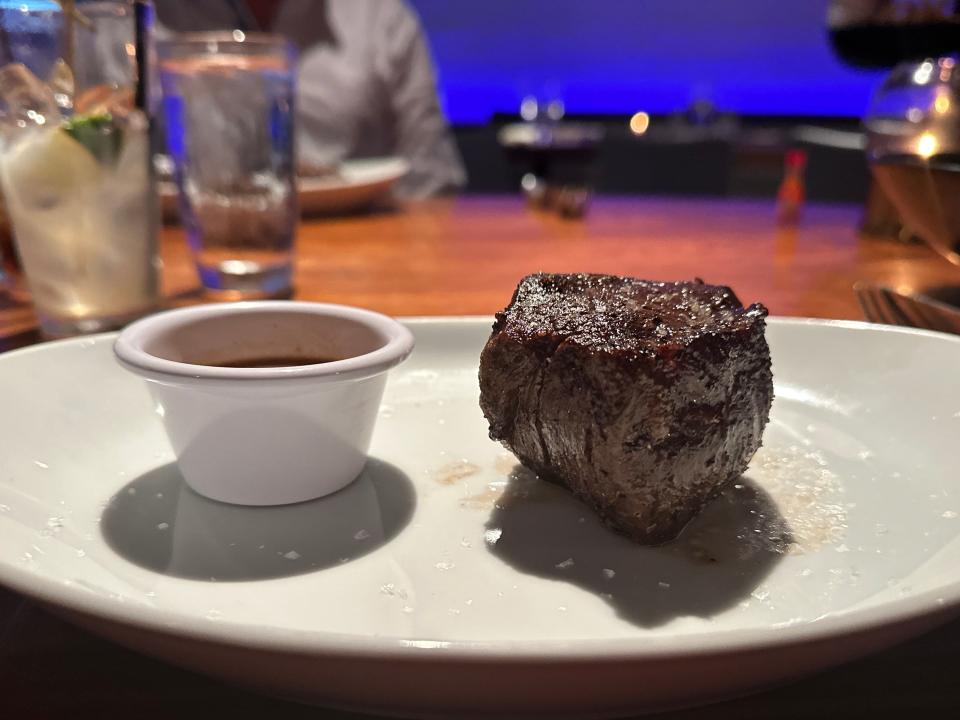 White plates with a piece of filet mignon next to a small white cup of a brown sauce with a brown table in the background