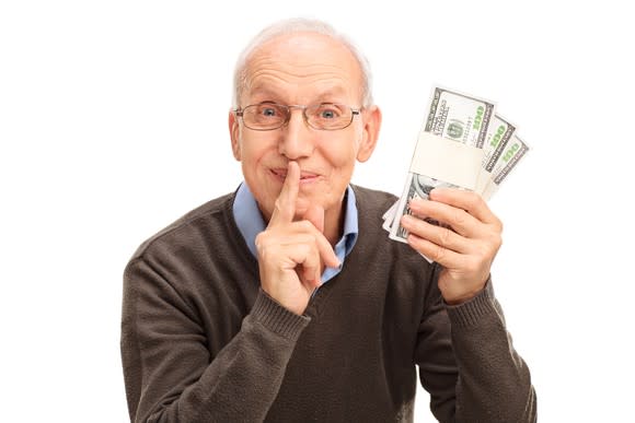 A senior man holds his finger to his lips while holding three stacks of banded hundred dollar bills.