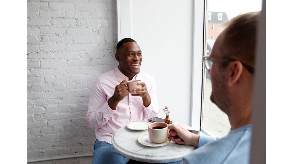 Man sitting at cafe table with a cup of coffee, smiling at another man