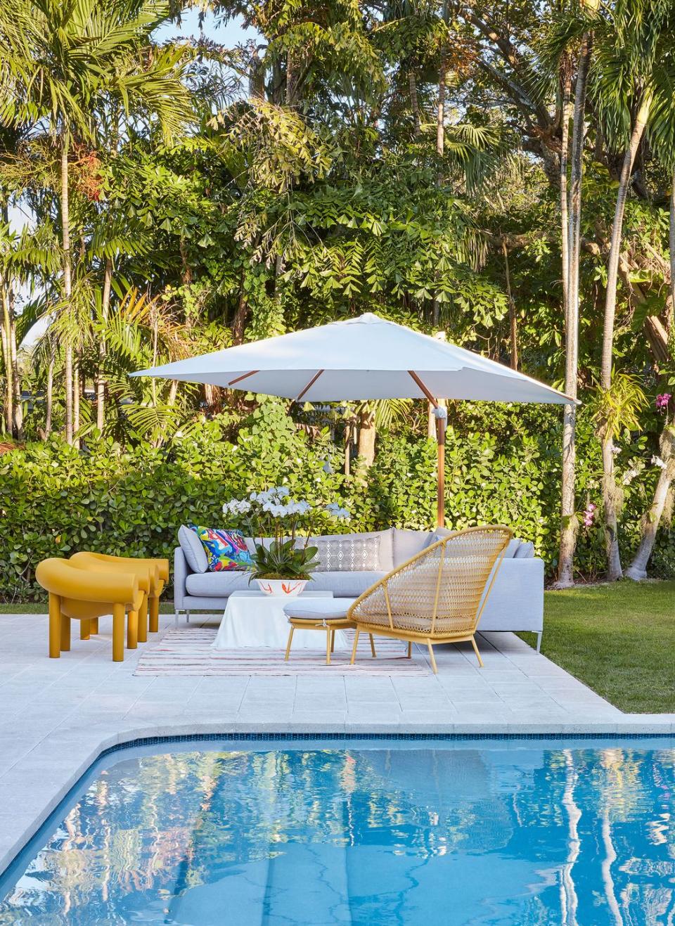 outside view of house by the pool where a seating area with a wraparound sofa table and several chairs and a large umbrella for shade