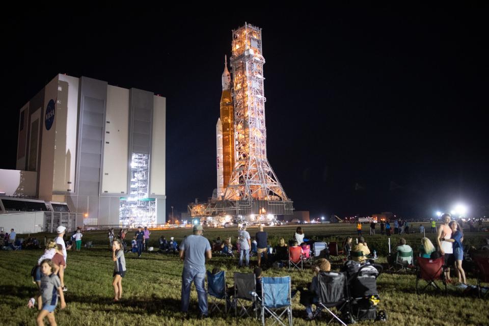 dozens of people standing in grass sitting in lawn chairs watch upright rocket roll out of tall building at night