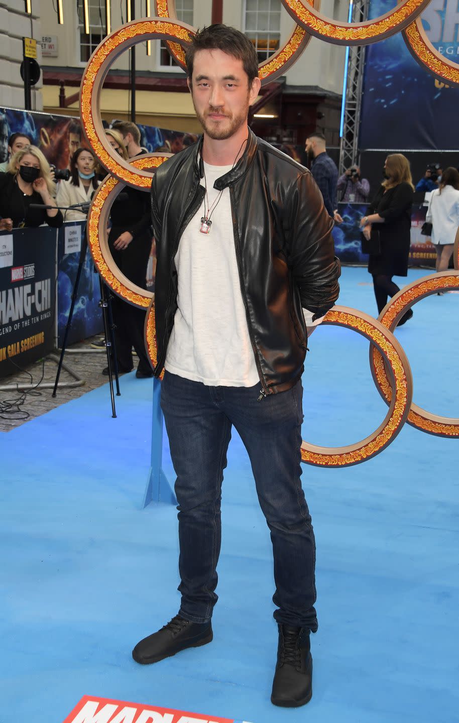 andrew koji, a man with dark hair and a beard wearing a white shirt and a black leather jacket, stands on a blue carpet in front of a sculpture of rings and smiles at the camera