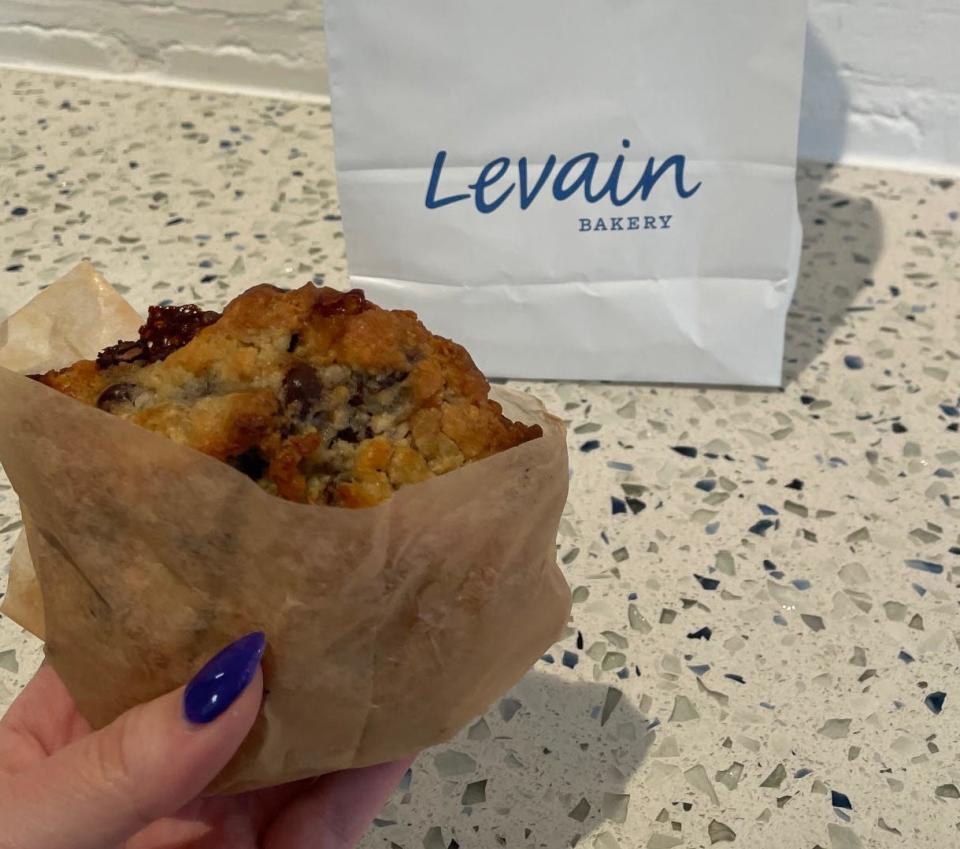 A person's hand holding a chocolate chip cookie in front of a Levain Bakery bag