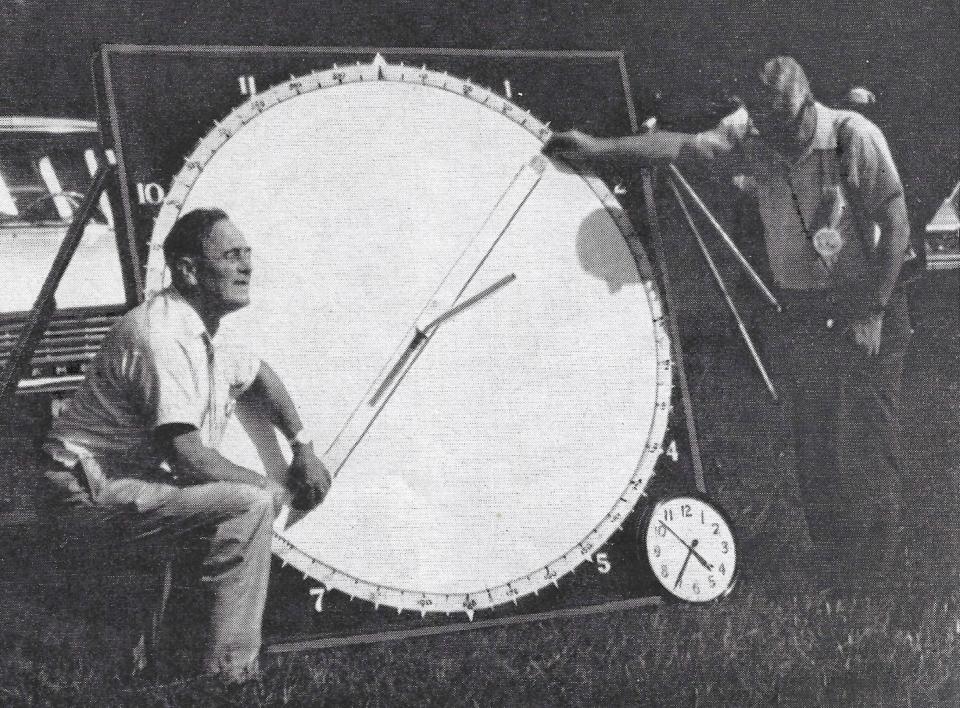 two men stand beside a large white circular screen that has a rod protruding from the center