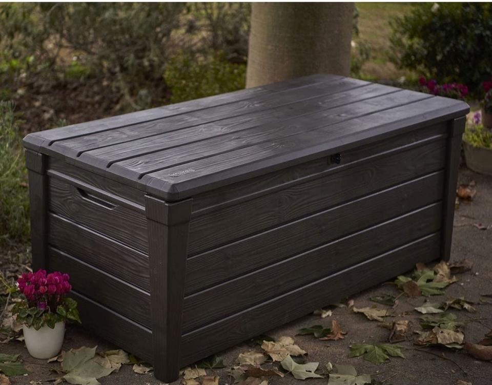 dark brown outdoor storage chest with a closed lid, placed on a patio next to a potted plant