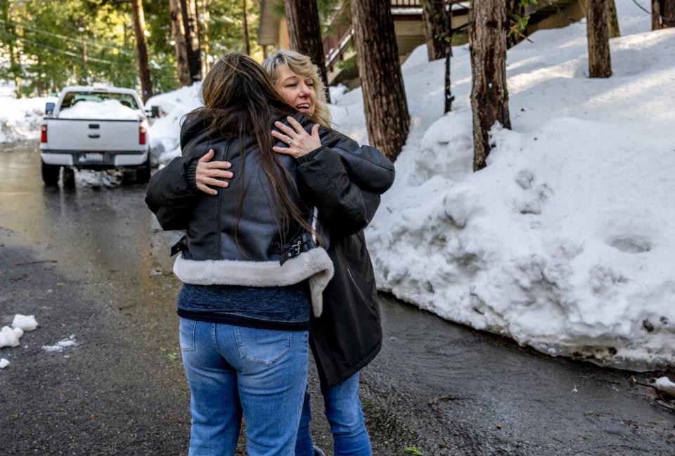 Two women in winter coats embrace on a road bordered by snow and pine trees.