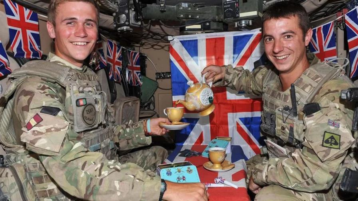 Two white people wearing camouflage uniforms are seated in front of British flags. The one on the left is holding a teacup on a saucer, while the one on the right is pouring tea from a teapot. . 