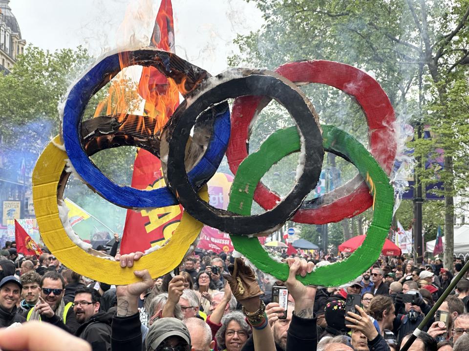 People holding up a burning Olympic rings symbol during a protest with a crowd in the background