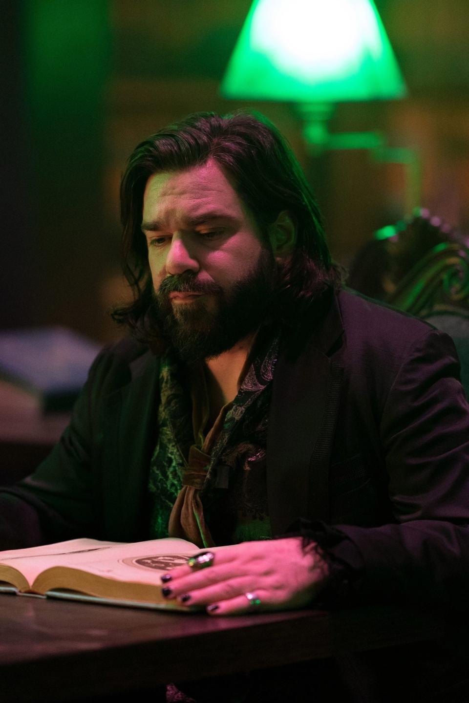 A bearded man in a dark, antique-style outfit reads a book under greenish lighting in a vintage library setting