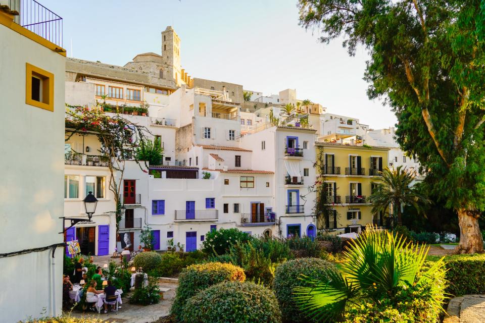A colorful street in Ibiza lined with vibrant homes and green trees and bushes.