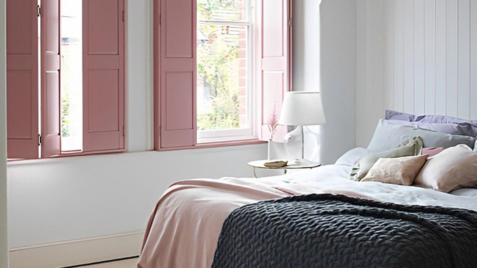  A white bedroom with pink wooden shutters, a pink throw and a glass nightstand light 