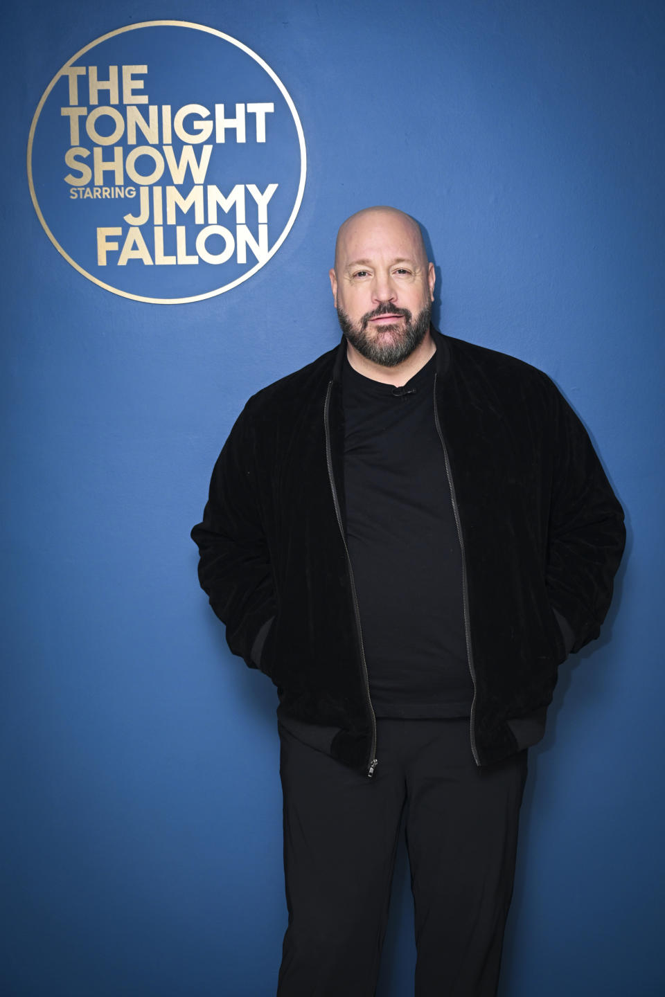 Kevin James stands in front of The Tonight Show Starring Jimmy Fallon sign, wearing a black outfit and jacket