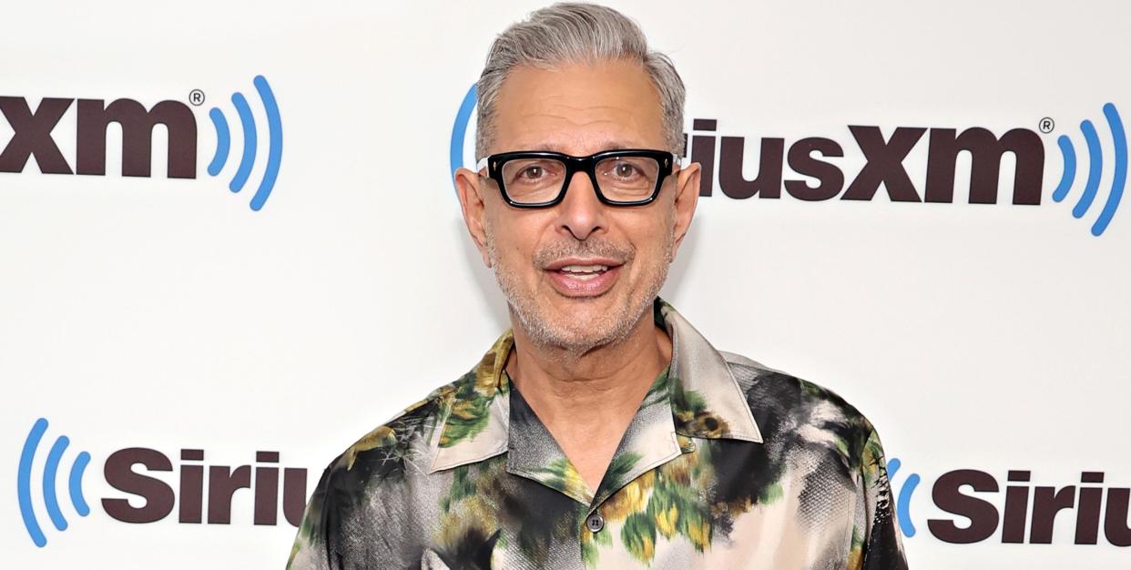 jeff goldblum, an older man stands looking at the camera with left hand in trouser pocket, greying hair, black thick rim glasses, wearing white and grey shirt with yellow flowers on and black trousers