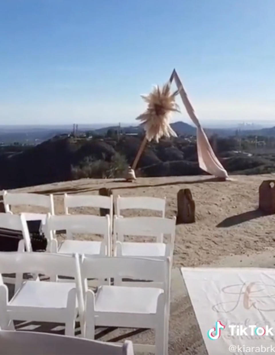 Picture of a wedding ceremony on a cliff, there is an arch with some dusty pink material and fluffy flower on it, white chairs are lined up on the left. 