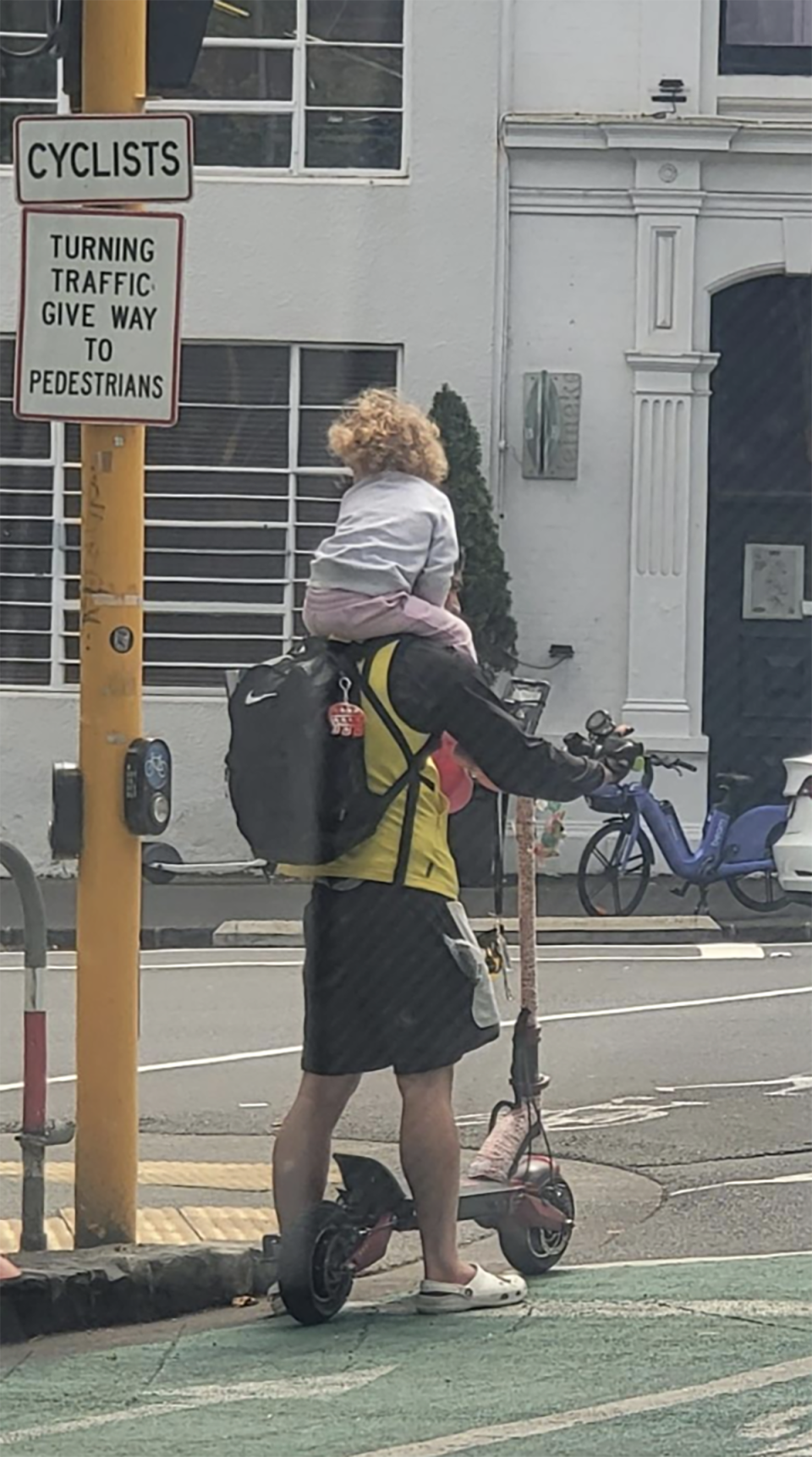 The e-scooter rider stopped at a red light on a road in the cyclist lane with a young girl not wearing a helmet sitting on his shoulders.